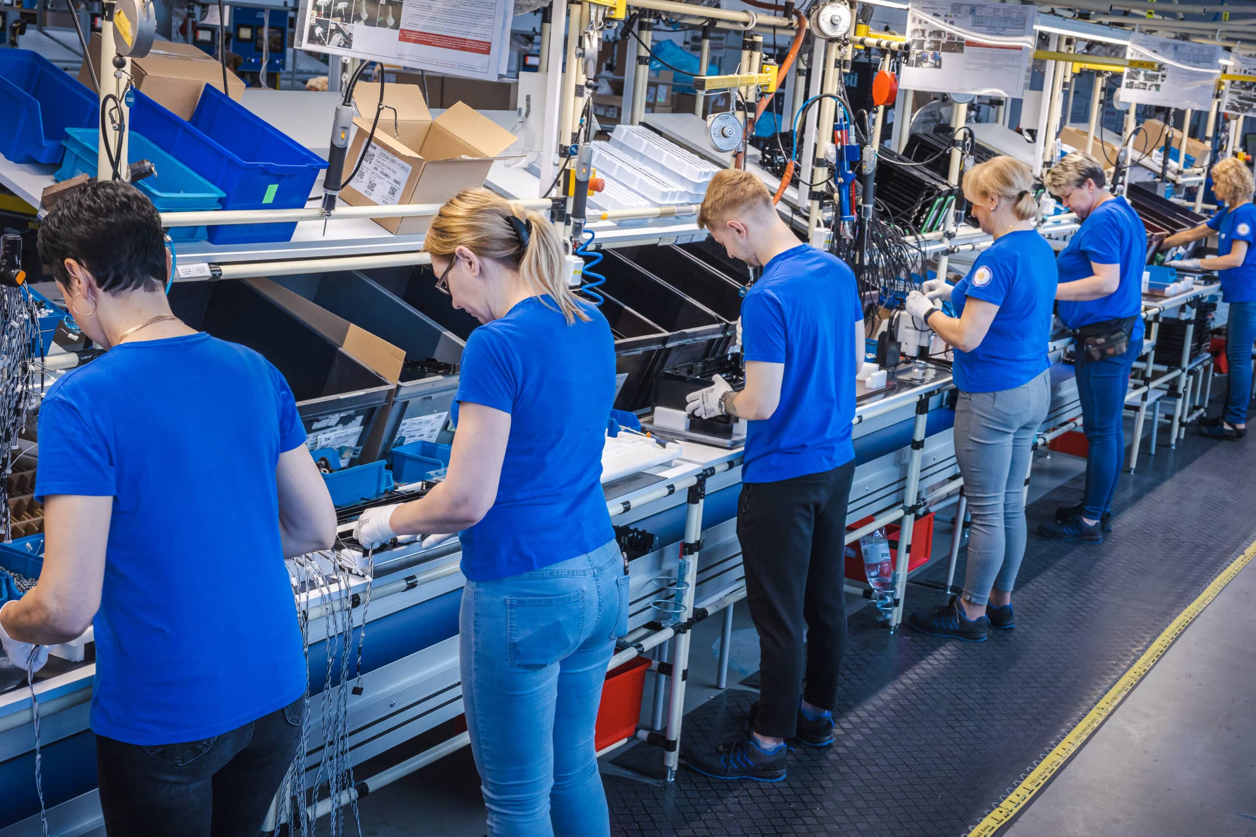 A group of workers wearing blue shirts assemble components along a production line in a brightly lit manufacturing facility. Each person focuses on individual tasks at organized workstations equipped with bins, tools, and wiring materials, illustrating teamwork, precision, and efficiency in a modern industrial environment.