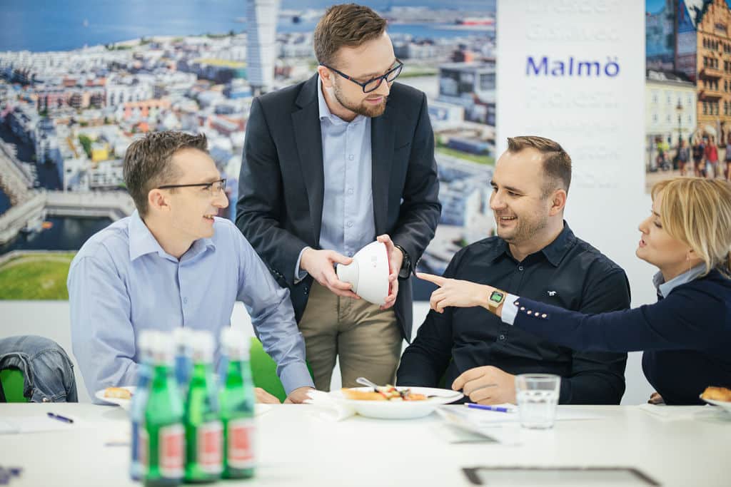 Four professionals sit around a conference table engaged in discussion, smiling and interacting. One person stands holding a white product prototype while the others look on attentively. The setting includes bottled water, notebooks, and a cityscape mural in the background, suggesting a collaborative business meeting or design review.