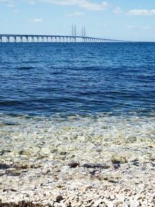 A long bridge stretches across a deep blue sea under a bright sky with scattered clouds. The foreground shows a rocky shoreline with clear, shallow water that transitions into darker blue as it deepens. The scene conveys calmness and the connection between land and sea.