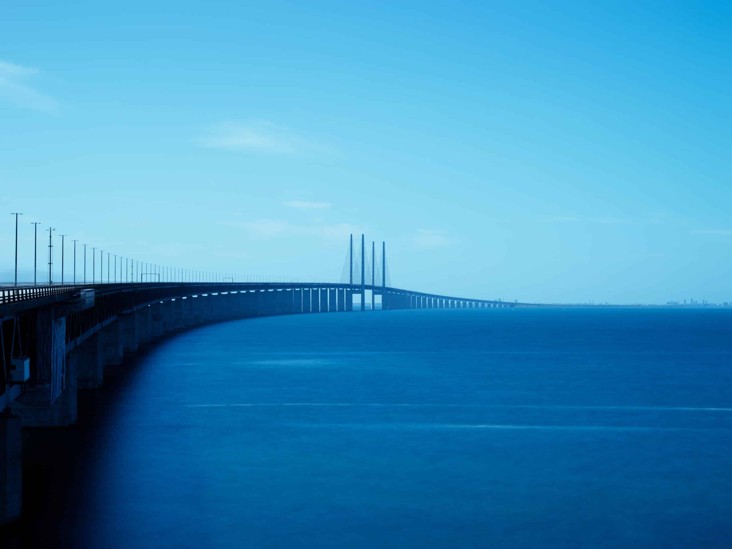 A long bridge stretches gracefully across a calm blue sea under a clear sky. The structure extends from the left side of the image into the distance. The overall scene conveys tranquility, symmetry, and vast open space
