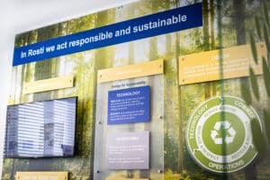 A sustainability wall display at Rosti featuring the message “In Rosti we act responsible and sustainable.” The board highlights sections on communication, awareness, technology, operations, and vision, emphasizing eco-friendly initiatives such as “Reduce & Reuse” and “Recycle & Recover.” The background shows a forest scene, reinforcing the company’s commitment to environmental responsibility