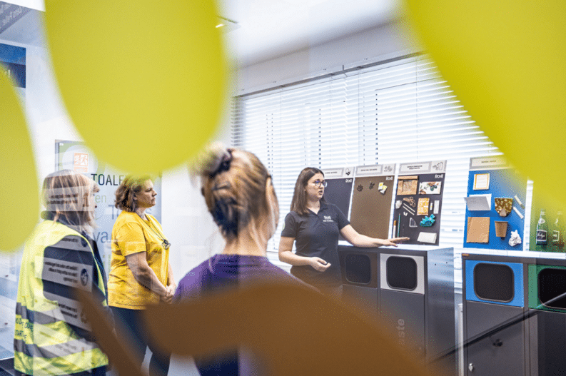 A woman gives a sustainability presentation to a small group, standing beside labeled recycling bins and display boards showing different waste materials. The attendees listen attentively in a brightly lit room with large windows, emphasizing education and awareness about proper recycling and waste sorting practices.