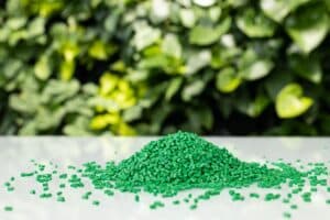 A small pile of green plastic pellets sits on a reflective white surface with more scattered around it. The background is softly blurred with lush green foliage, creating a natural, eco-friendly contrast to the synthetic material in the foreground.