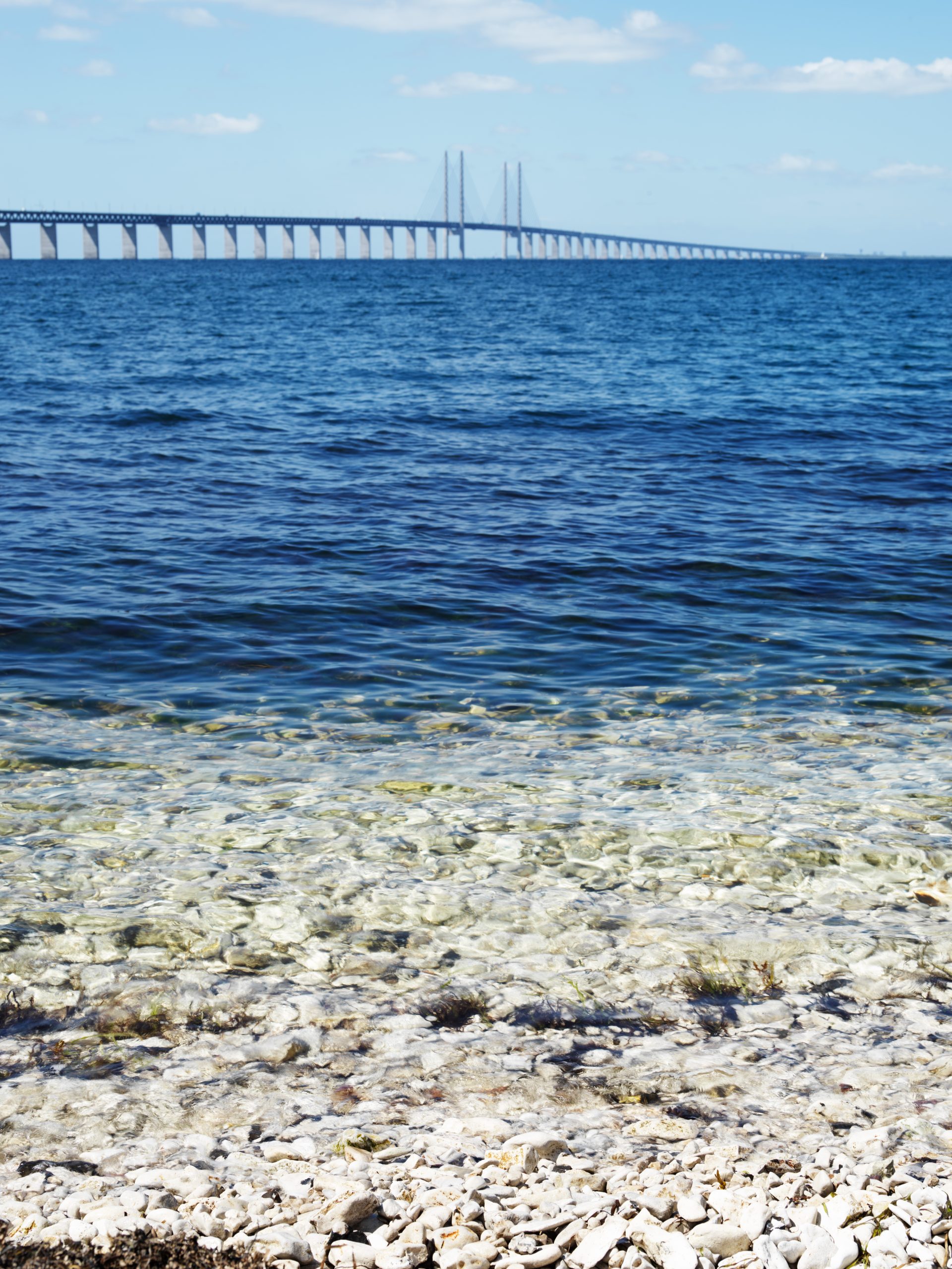 Parlak gökyüzünde dağılmış bulutların altında derin, mavi bir denizin üzerinden uzun bir köprü uzanıyor. Ön planda berrak ve sığ suyun derinleşerek koyulaştığı, kayalık bir kıyı görünüyor. Sahne huzuru ve kara ile deniz arasındaki bağlantıyı yansıtıyor.